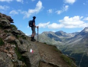 Eine Frau mit Rucksack und Wanderstöcken auf einem exponierten Wanderweg mit Blick auf ein weitläufiges Bergpanorama