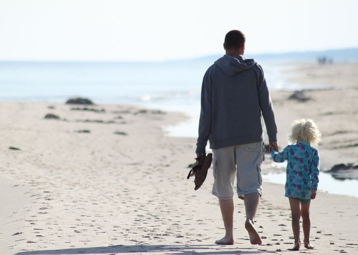 Kleinkind mit blonsen Locken an der Hand des Vaters am Strand