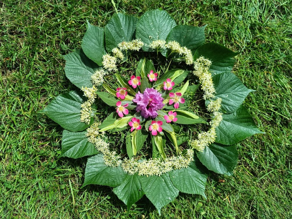 Ein Mandala aus grünen, roten und weißen Blüten und Blättern auf einer Wiese.